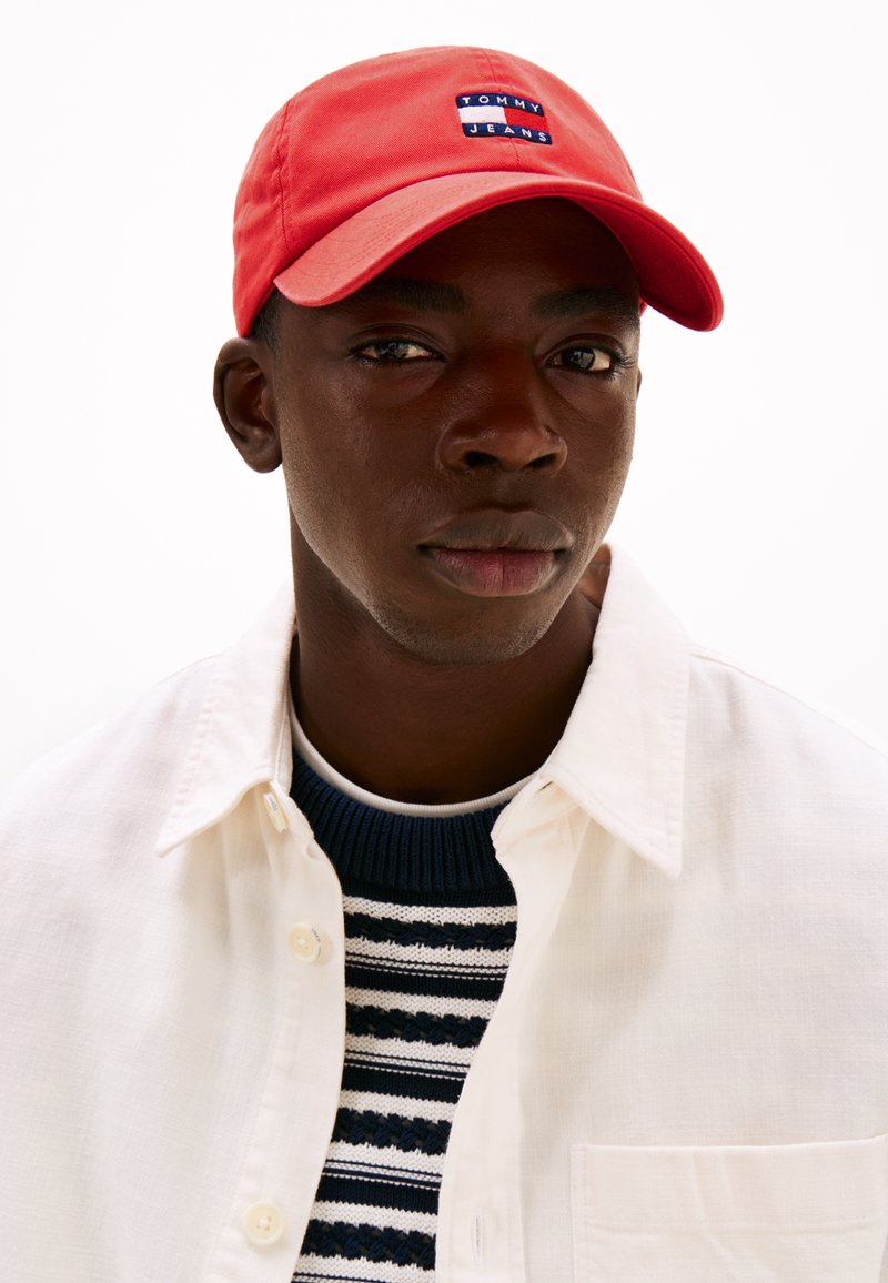 Rød cap med logo-patch, båret med en hvid skjorte og en navy stribet sweater. Nærbillede, der fremhæver ansigt og tøj.