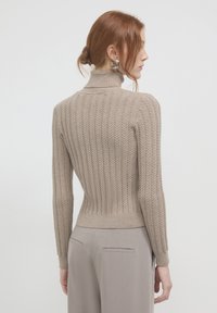 Frau mit rotem Haar in einem niedrigen Dutt, die große silberne Creolen trägt, einen beigen strukturierten Rollkragenpullover und hellgraue Hosen an hat und seitlich steht.