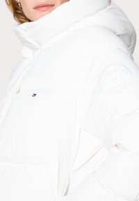 Biała kurtka puchowa z wysokim kołnierzem, teksturowaną powierzchnią, elastycznym dołem i haftowanym logo na rękawie. Materiał wydaje się miękki i lekki.