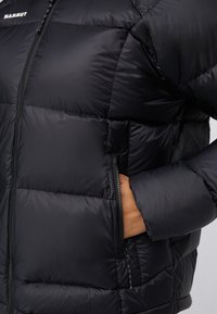 Veste en duvet noire avec un design matelassé, des poches zippées et une texture lisse. Logo affiché près du col.