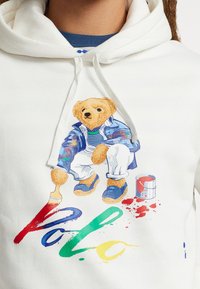 Vit hoodie med en färgglad björn grafik, iklädd en blå jacka och blå skor, håller en pensel med färgfläckar och "Polo" text.