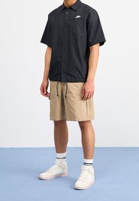 Svart kortärmad skjorta med knappar och bröstficka, prydd med en vit logotyp, tillsammans med beige shorts och vita höga sneakers.