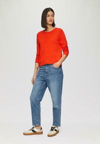 Rode gebreide cardigan met lange mouwen en knopen aan de voorkant, gecombineerd met lichtblauwe recht-toe-rechtdoor jeans en zwart-witte sneakers.