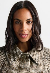 Manteau à motif à carreaux marron et crème avec un col rond et un grand bouton. Le tissu semble texturé et chaud.