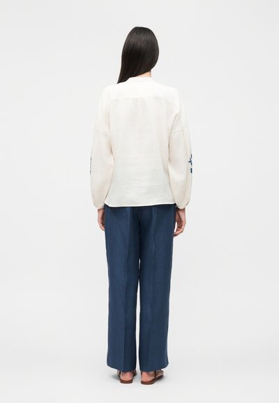 Γυναίκα με μακριά σκούρα μαλλιά στέκεται με την πλάτη, φορώντας φαρδιά λευκή μπλούζα με μπλε κεντήματα στα μανίκια και φαρδιές μπλε παντελόνες με σανδάλια.