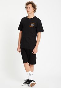 T-shirt en coton noir avec un graphique orange et blanc sur la poitrine gauche, associé à un short noir, des chaussettes logo blanches et des baskets noires.