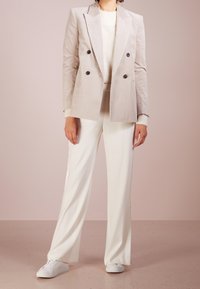 Helles beiges Kordblazer im Doppelreiher-Stil mit schwarzen Knöpfen, kombiniert mit einem cremefarbenen Strickpullover und weißen Hosen, abgerundet mit weißen Sportschuhen.