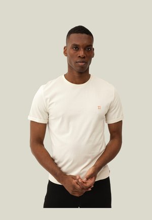 Camiseta de manga corta color crema, fabricada en algodón, con un pequeño emblema naranja "II" en el pecho, con una silueta ajustada y mangas enrolladas.