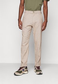 Light beige trousers with a textured finish, featuring a single button waistband and side pockets, paired with multicolored athletic shoes.
