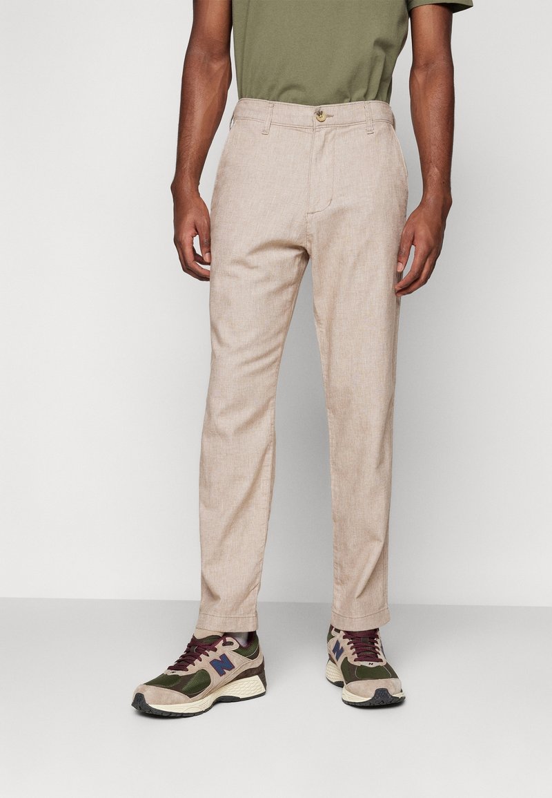 Light beige trousers with a textured finish, featuring a single button waistband and side pockets, paired with multicolored athletic shoes.