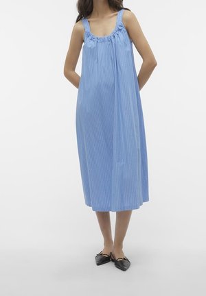 Woman wearing a loose, sleeveless blue midi dress with gathered neckline and black pointed-toe flats, standing against a white background.