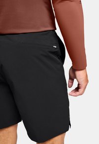 Shorts de sport noirs avec une texture lisse, comprenant une poche latérale et une petite étiquette de marque à l'arrière, associés à un haut marron à manches longues.