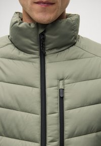 Ανοιχτό ελαιόλαδο πράσινο padded μπουφάν με ψηλό γιακά, διαθέτοντας μαύρους φερμουάρ και πλαϊνή τσέπη. Το υλικό φαίνεται ομαλό και συνθετικό.