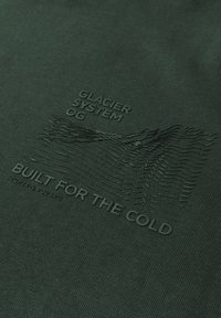 Dunkelgrüner Stoff mit geprägtem Text: "GLACIER SYSTEM OG" und "BUILT FOR THE COLD", darüber befinden sich strukturierte Wellenmuster.