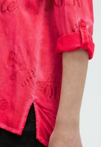 Rotes Langarmhemd mit hochgerolltem Ärmel, mit bedruckten Textmustern und seitlichem Schlitz, getragen mit schwarzen Hosen.