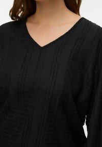 Femme vêtue d'un haut noir texturé à manches longues et encolure en V, dévoilant le cou et le haut de la poitrine, avec des cheveux marron lâches.