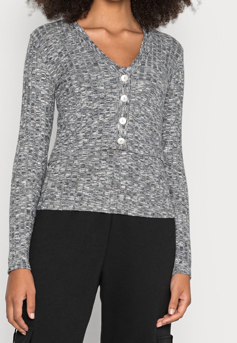 Femme portant un haut à manches longues gris texturé avec un col en V et quatre boutons blancs sur le devant, associé à un pantalon noir.