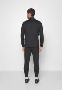 Schwarzes Sportoberteil mit langen Ärmeln und hohem Kragen, kombiniert mit passenden, eng anliegenden Hosen. Das Material scheint leicht und glatt zu sein.