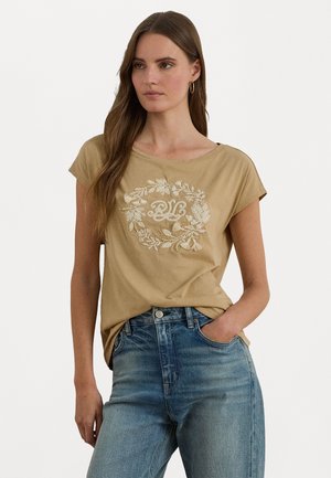 T-shirt beige a maniche corte in cotone con un motivo circolare floreale ricamato in bianco, abbinato a jeans in denim azzurri.