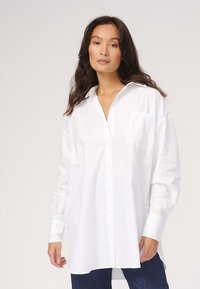 Hvid skjorte med knapper, afslappet pasform, lange ærmer og en enkelt brystlomme, lavet af glat stof. Bæres med blå denim.