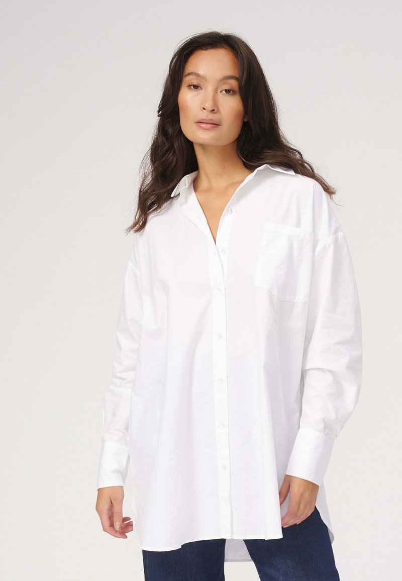 Hvid skjorte med knapper, afslappet pasform, lange ærmer og en enkelt brystlomme, lavet af glat stof. Bæres med blå denim.
