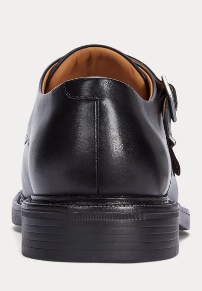 Sapatos pretos de couro com acabamento liso, com formato arredondado, salto pequeno e detalhe de fivela lateral. O interior é castanho.