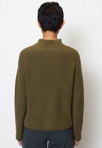 Eine Person trägt einen olivgrünen, gerippten Strickpullover und steht mit dem Rücken zu einem schlichten weißen Hintergrund.