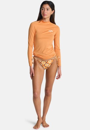 Donna in piedi a piedi nudi che indossa una maglia protettiva arancione a maniche lunghe e slip bikini con motivo floreale su sfondo semplice.