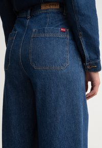Vaqueros de mezclilla en azul oscuro con una cintura alta, que presentan dos bolsillos traseros, costuras sutiles y una etiqueta de logo en el bolsillo derecho.