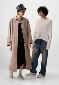 Deux personnes debout, l'une portant un trench-coat beige, une robe noire, un chapeau seau noir et des baskets lavande ; l'autre un pull en maille blanc, un jean bleu et des baskets.