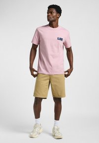 Camiseta de algodón rosa con un pequeño logo en azul marino "LEE" en el pecho izquierdo, combinada con pantalones cortos beige y zapatillas de crema.