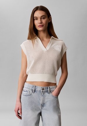 Jeune femme portant un top en maille blanche sans manches avec col et un jean taille haute bleu clair, debout, une main dans la poche, sur fond gris.