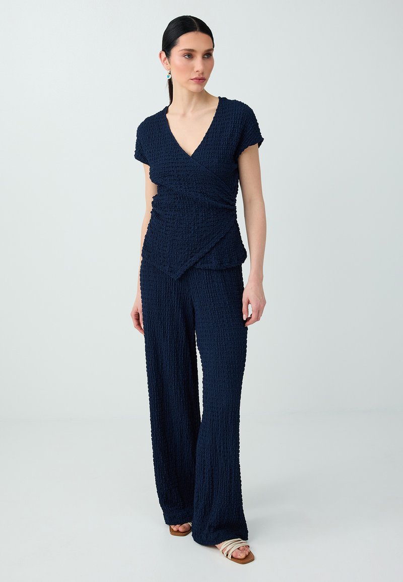 Femme debout, portant un haut portefeuille bleu marine texturé et un pantalon large assorti, avec des sandales blanches, sur un fond clair et uni.
