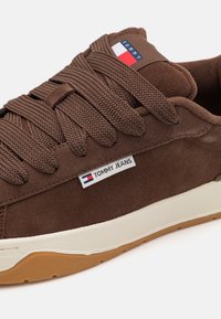 Brune semskede sneakers med teksturerte snørebånd, hvit mellomsåle og brun gummisåle. Har et "Tommy Jeans"-logo på siden.