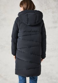 Abrigo de plumas negro con capucha, que presenta una textura acolchada y costuras diagonales. De largo, se lleva con jeans azules.