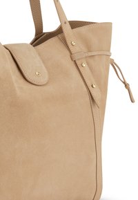 Sac fourre-tout en daim de couleur beige clair avec un rabat, ferrures dorées et couture décorative. Comprend des liens latéraux ajustables pour la forme.