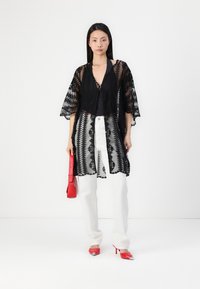 Svart spetskimono med geometriska mönster, korta ärmar och scallopade kanter. Kombinerad med vita byxor och röda accessoarer för kontrast.