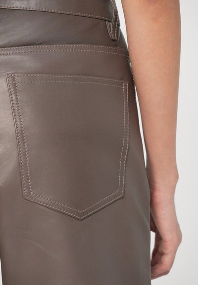 Close-up van de heup en arm van een persoon die een bruin leren broek draagt met zichtbare gestikte achterzak en schijndetails.