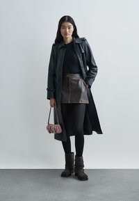 Trench coat in pelle nera sopra un top nero e mini gonna in pelle marrone. Porta una piccola borsa rosa e indossa stivaletti marroni.