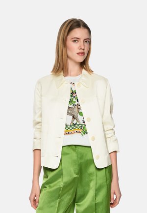 Frau trägt eine weiße Jacke mit Knöpfen über einem bedruckten Hemd mit Leopardenmuster, kombiniert mit glänzenden grünen, weit geschnittenen Hosen, stehend vor weißem Hintergrund.