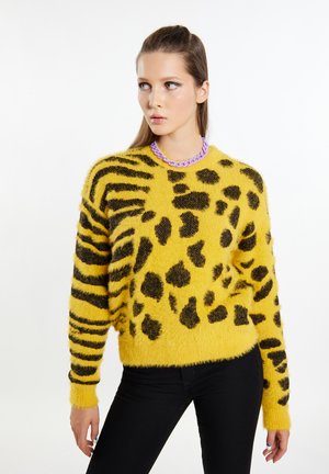 Geel pluizig trui met zwarte luipaard- en tijgerstrepen, ronde halslijn en een contrasterende paars kleurige ketting. Zwarte jeans maken de outfit compleet.