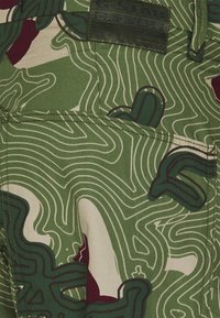 Tecido verde com um padrão topográfico em bege e bordeaux, apresentando detalhes de costura e uma etiqueta de couro G-Star Originals.