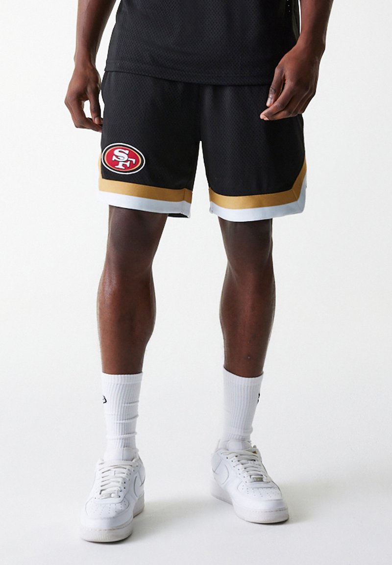 Svart mesh basketballshorts med rødt og hvitt logo, gull- og hvite striper i kanten, brukt med hvite sokker og sneakers.