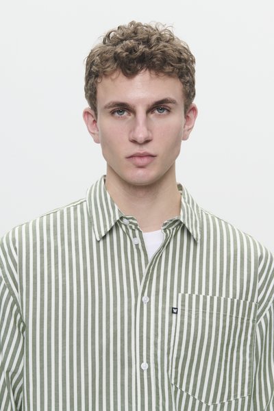 Giovane con capelli ricci che indossa una camicia a righe verticali verde e bianca con taschino, davanti a uno sfondo neutro.