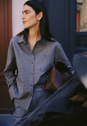 Jeune femme aux longs cheveux foncés porte une chemise à carreaux boutonnée et un pantalon gris, tenant un sac en cuir marron, debout à l'extérieur.