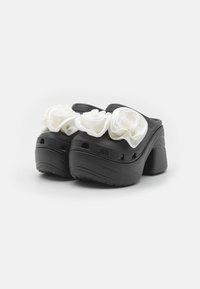 Black clogs with a chunky heel and a textured surface, featuring large white satin rose accents on the front.