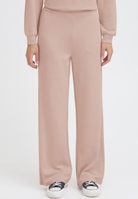 Hellrosa Hosen mit weitem Bein aus einem weichen Stoff. Sie zeichnen sich durch eine glatte Textur und einen elastischen Bund aus und werden mit schwarzen Sneakers kombiniert.