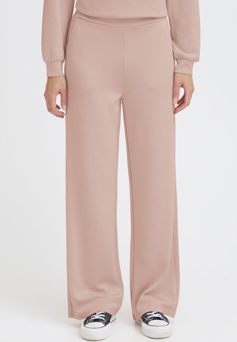 Hellrosa Hosen mit weitem Bein aus einem weichen Stoff. Sie zeichnen sich durch eine glatte Textur und einen elastischen Bund aus und werden mit schwarzen Sneakers kombiniert.
