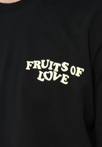 T-shirt preta de algodão com texto amarelo curvado que diz "FRUITS OF LOVE" no lado esquerdo. Textura suave, design clássico com gola redonda.