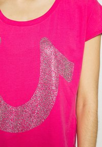 Camisa de manga corta de color rosa brillante con un gran diseño de herradura engastado en strass plateados en el pecho, usada por una persona de piel clara.